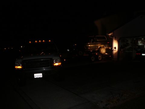 1991 Ford Bronco - A white Ford F-250 Super Duty pickup truck is parked at night in front of a garage. A dark-colored Ford Bronco is loaded onto a trailer beh…