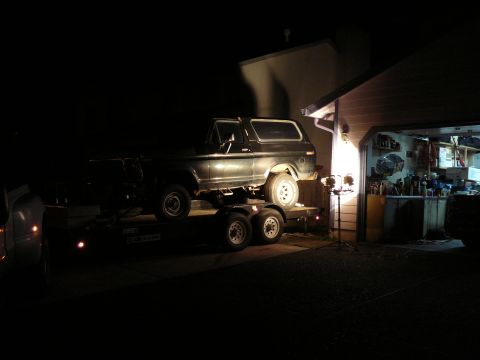 1991 Ford Bronco - A dark-colored SUV is parked on a trailer at night, illuminated by spotlights. The vehicle is positioned in front of an open garage filled…
