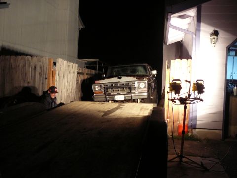 1991 Ford Bronco - A man in a baseball cap and dark jacket leans over a wooden ramp next to a classic Ford pickup truck at night. Two portable work lights ill…