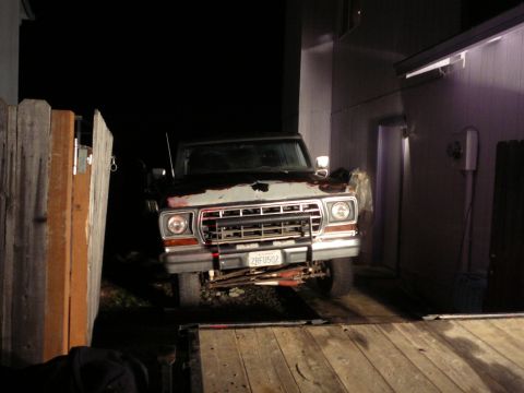 1991 Ford Bronco - The front view of a vintage Ford pickup truck is shown at night, parked next to a wooden fence and a building. The truck has a rusted hood…