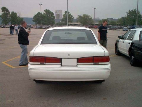 2000 Ford Crown Victoria - Straight-on rear view of a white full-size four-door sedan in a parking lot, showing full-width red tail lamps, a blank white license plate…