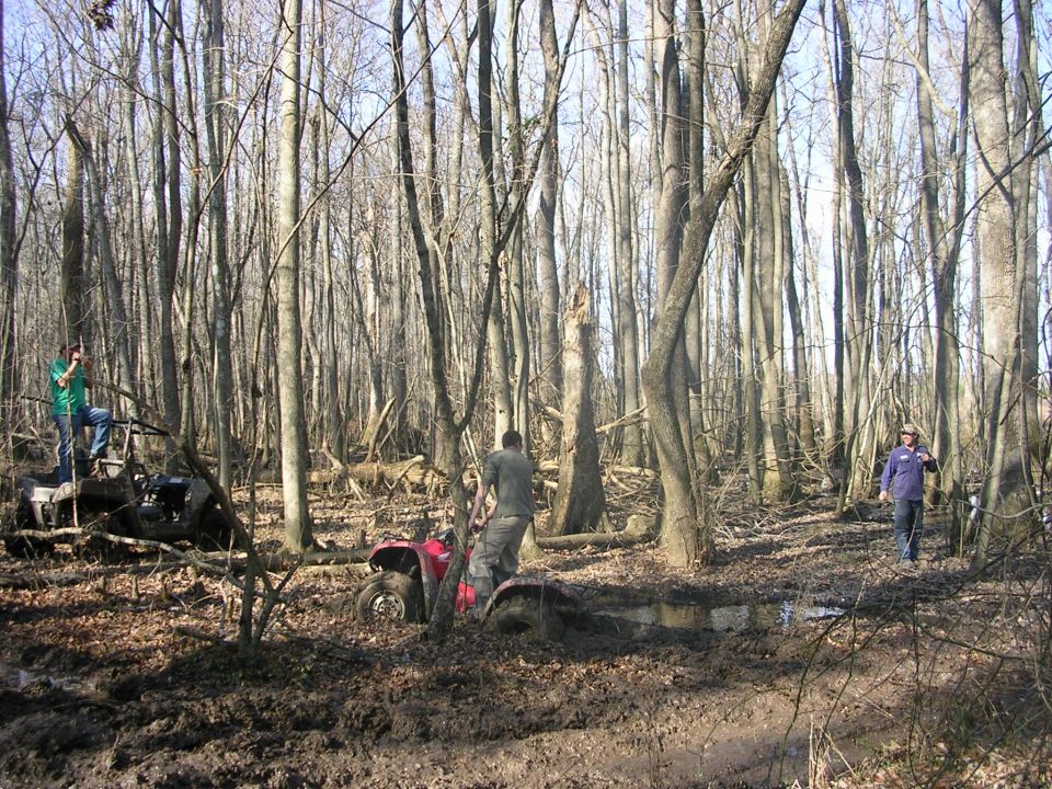 Red ATV stuck axle-deep in a mud hole in leafless winter woods with riders nearby