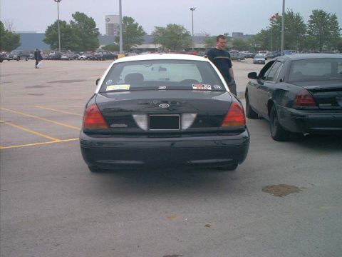 2000 Ford Crown Victoria - Straight-on rear view of a black Ford Crown Victoria sedan with a white roof and blacked-out plate, parked in a large mall lot with event d…