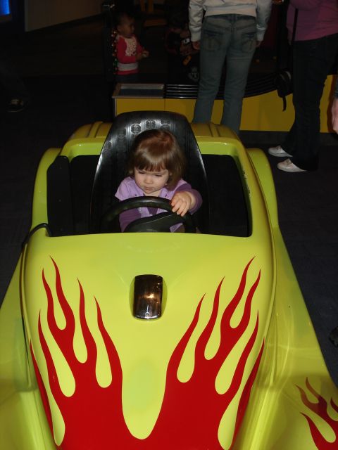 2000 Ford Crown Victoria - A young girl sits in the driver's seat of a bright yellow toy car with red flame decals. The car has a black interior and a chrome detail o…