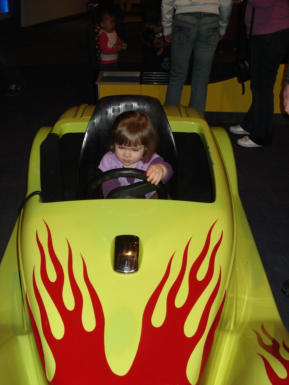 Young girl in a yellow toy car with red flame decals.