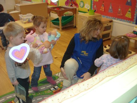 2000 Ford Crown Victoria - A woman in a blue apron interacts with children in a classroom setting. One child holds several dolls, while another child holds a heart-sh…