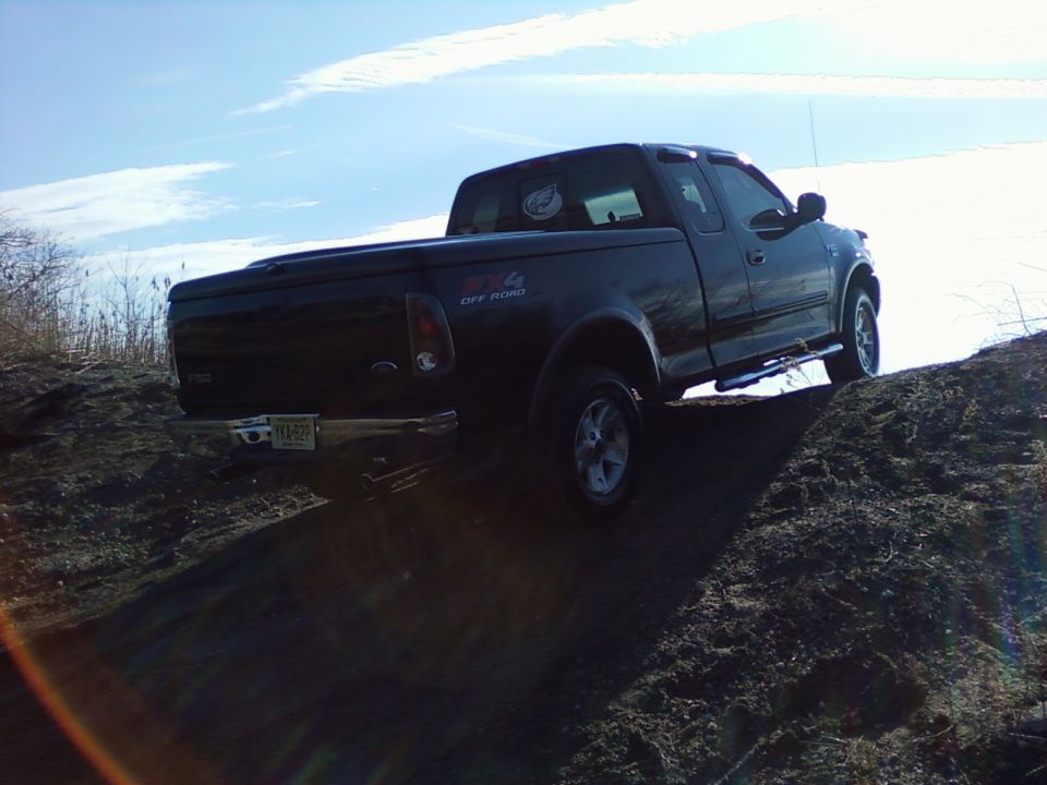 Black 2002 Ford F-150 pickup truck driving up a dirt hill, rear-side view.
