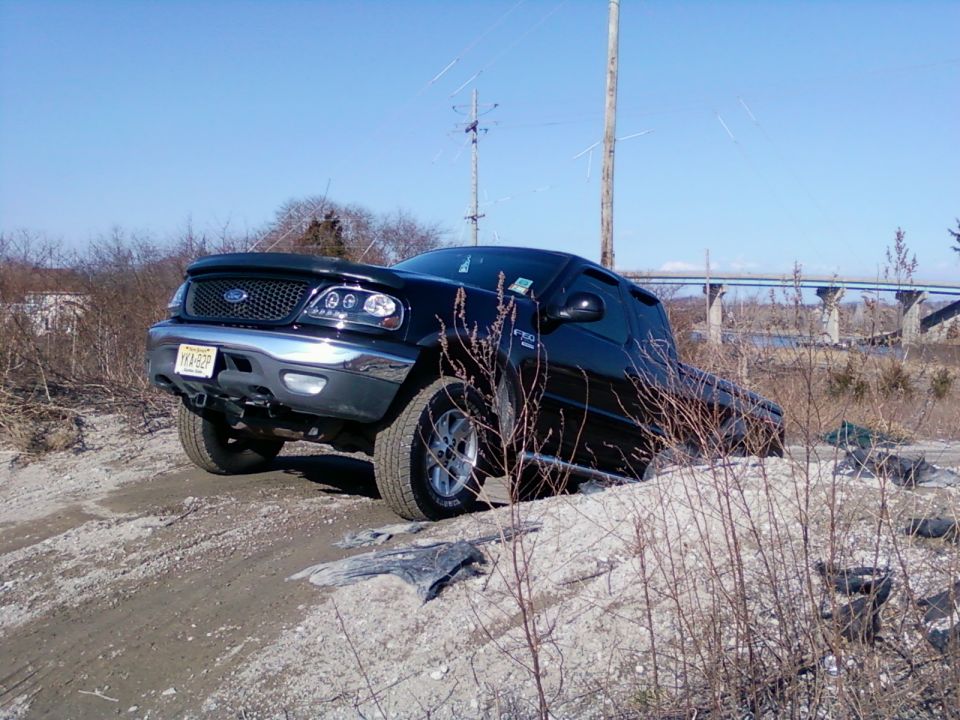 Black Ford F-150 pickup truck on a dirt incline with dry brush and a bridge in the background.