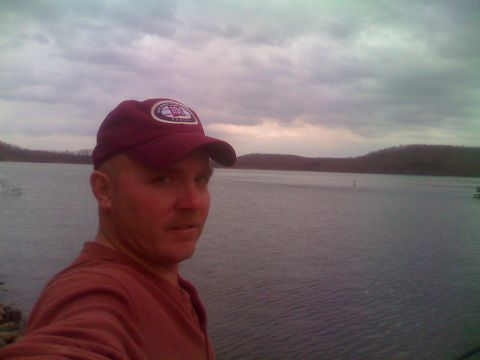 1996 Ford Bronco - A man wearing a maroon baseball cap and a maroon long-sleeved shirt stands in front of a large body of water under a cloudy sky.