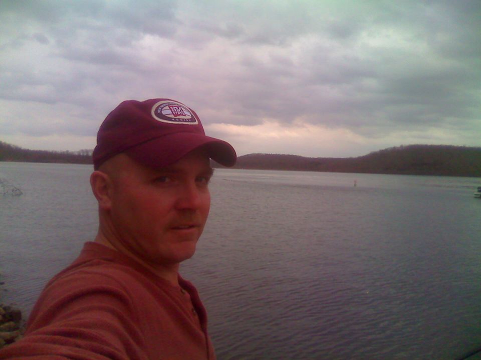 Man in a maroon baseball cap and shirt by a lake.