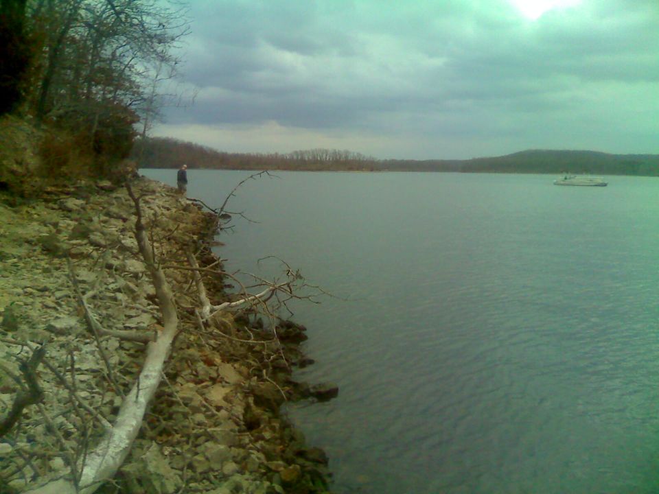 Person on rocky shore next to a lake with a boat in the distance.