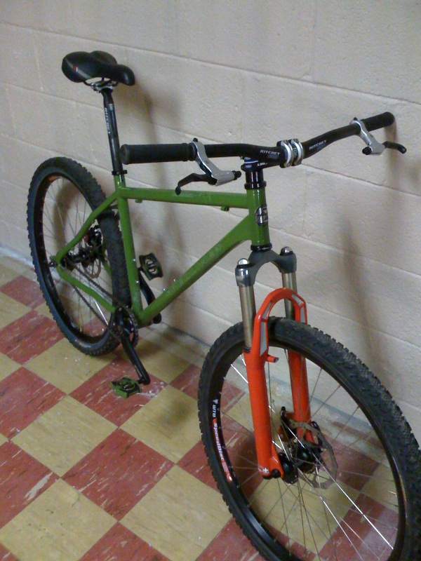 Green hardtail mountain bike with orange suspension fork leaning against a cinder block wall