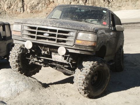 1995 Ford Bronco - A muddy 1995 Ford Bronco is shown at a low front 3/4 angle, positioned on a dirt path with a large rock on its left. The vehicle has large…