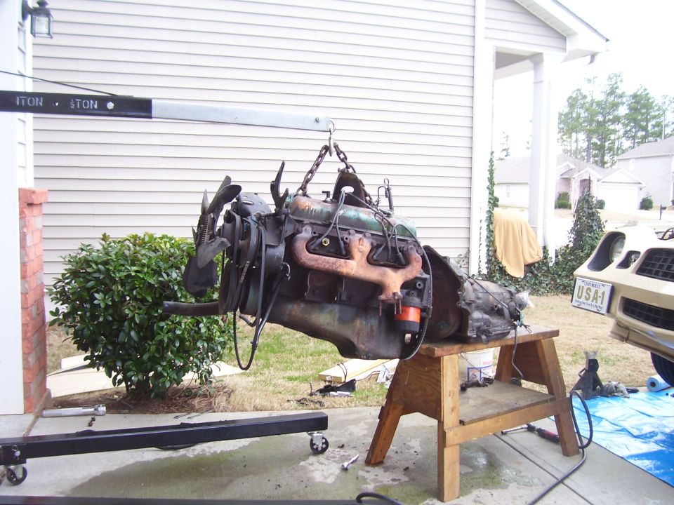 Engine and transmission assembly hanging from hoist, with a 1979 Camaro visible.