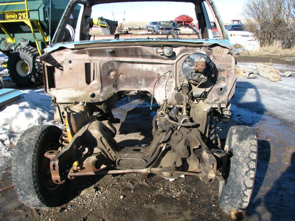Front view of a rusted pickup truck chassis with engine and suspension parts, outdoors.