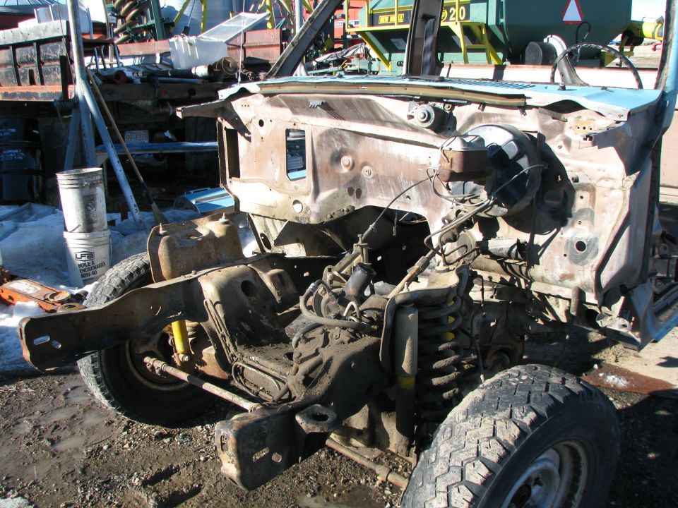 Rusted front chassis and engine bay of a disassembled blue pickup truck in a junkyard.