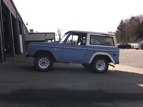 1996 Ford Bronco - A light blue first-generation Ford Bronco with a white hardtop is parked in front of a building with open bays. The vehicle is viewed from…