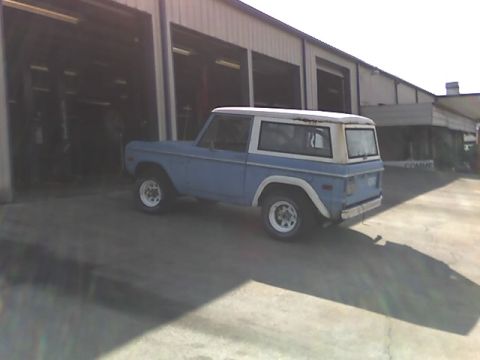 1996 Ford Bronco - A light blue Ford Bronco with a white hardtop is parked outside a garage with open bay doors. The vehicle is viewed from the rear three-qua…