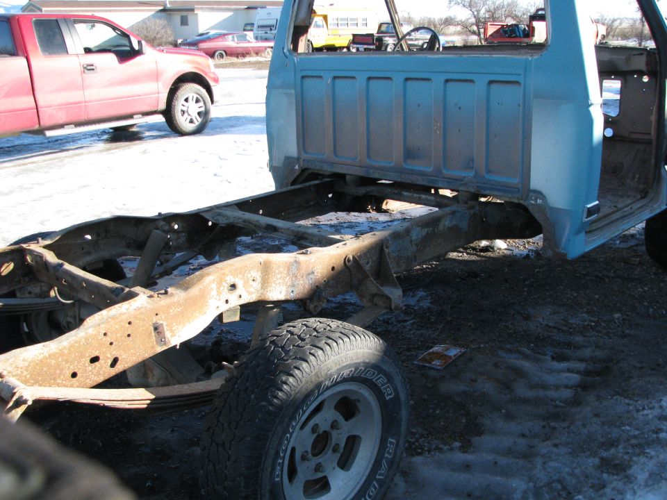Rusted chassis and frame of a light blue pickup truck, with a red pickup in the background.