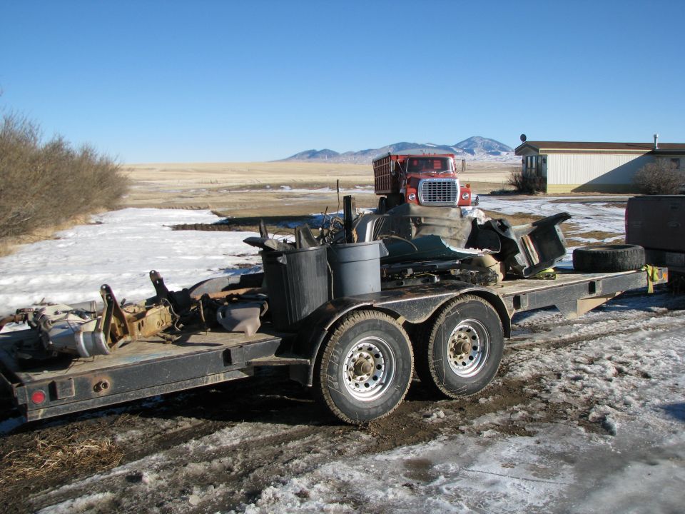Flatbed trailer loaded with truck parts, including a red truck cab, in a snowy, rural setting.