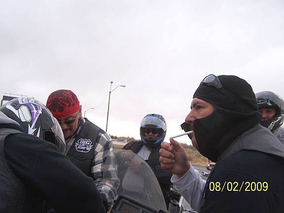 Riders in helmets and balaclavas gathered around a motorcycle at a cold roadside stop