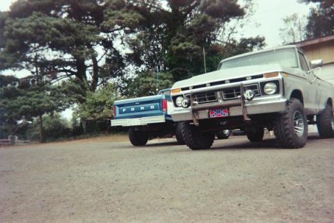 1977 Ford F-150 - Two pickup trucks are parked on a gravel surface in front of trees and a building. The truck in the foreground is a light-colored Ford with…