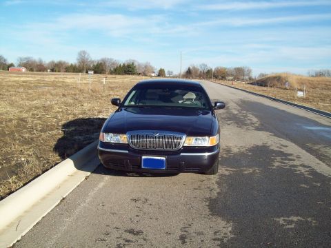 1999 Mercury Grand Marquis - A dark blue Mercury Grand Marquis is parked on the side of a wet asphalt road with a dry, grassy field to the left. The car is facing the c…