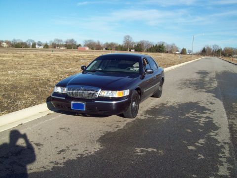 1999 Mercury Grand Marquis - A dark blue Mercury Grand Marquis sedan is parked on the side of an asphalt road. The car is facing slightly left, with a field and trees i…