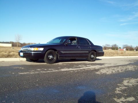 1999 Mercury Grand Marquis - A dark-colored sedan is parked on the side of a paved road with a dry, grassy field and scattered trees in the background under a clear blu…