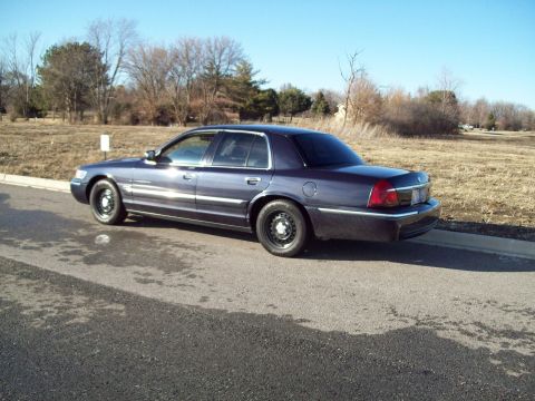 1999 Mercury Grand Marquis - A dark blue four-door sedan is parked on the side of a road with a dry grassy field and trees in the background. The car is viewed from the…