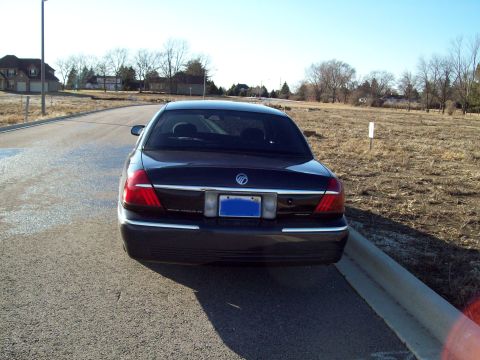 1999 Mercury Grand Marquis - The rear view of a dark colored sedan parked on the side of a road. The car has a blue license plate and chrome trim around the taillights…