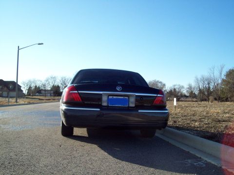 1999 Mercury Grand Marquis - The rear view of a dark-colored sedan parked on an asphalt road next to a curb. The car has a blue license plate and chrome trim around the…