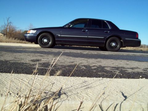 1999 Mercury Grand Marquis - A dark blue Mercury Grand Marquis sedan is parked on the side of a road with a clear blue sky in the background. The car is viewed from the…