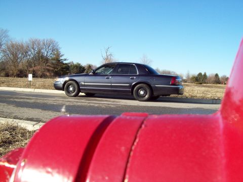 1999 Mercury Grand Marquis - A dark blue Mercury Grand Marquis sedan is parked on the side of a road with a bright blue sky overhead. The foreground is dominated by a b…