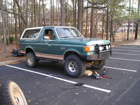 1989 Ford Bronco - Green Ford Bronco with white hardtop and tan lower body sits in a wooded parking lot mid-job, front passenger wheel removed and the hub res…