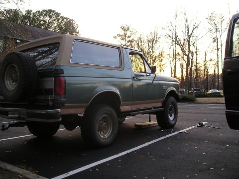 1989 Ford Bronco - Driver-side rear three-quarter view of a green and tan Ford Bronco with a tan removable top and swing-away spare carrier, sitting on a jack…