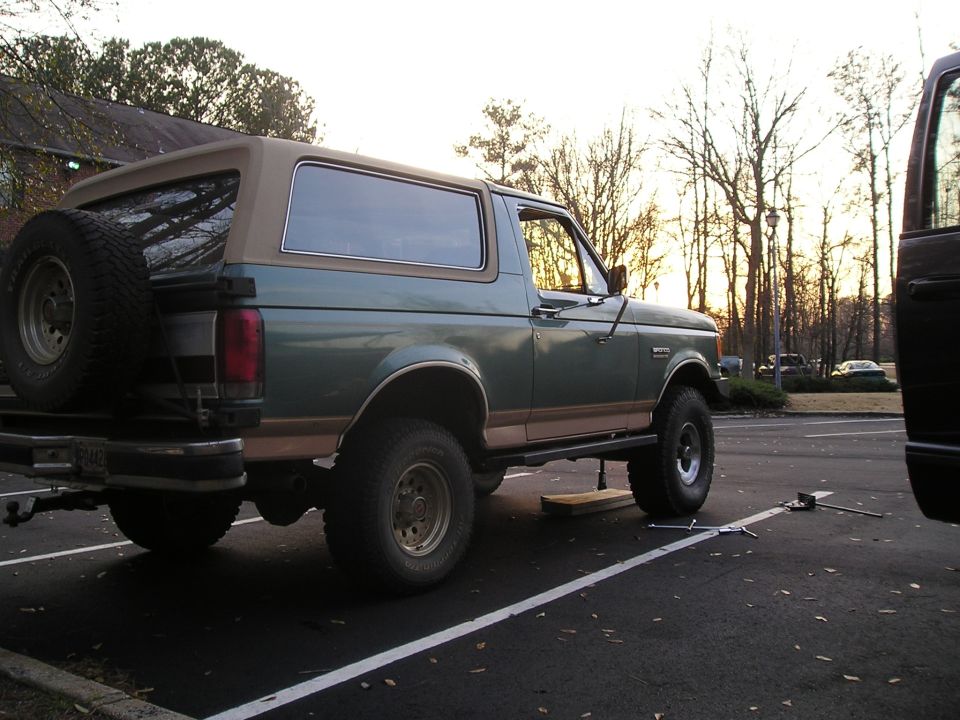 Green and tan Ford Bronco jacked up in a parking lot at sunset with tools nearby