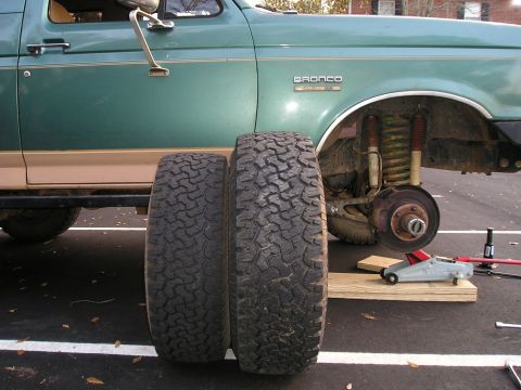 1989 Ford Bronco - A green and tan Ford Bronco sits jacked up in a parking lot with its front wheel removed, exposing the coil spring, shock, front hub and du…
