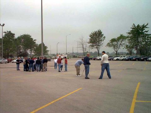 2000 Ford Crown Victoria - A gathering of people in a large parking lot, with several cars visible in the background under an overcast sky.