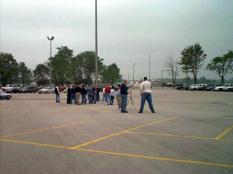 2000 Ford Crown Victoria - A group of people stand in a parking lot with cars visible in the background under an overcast sky.