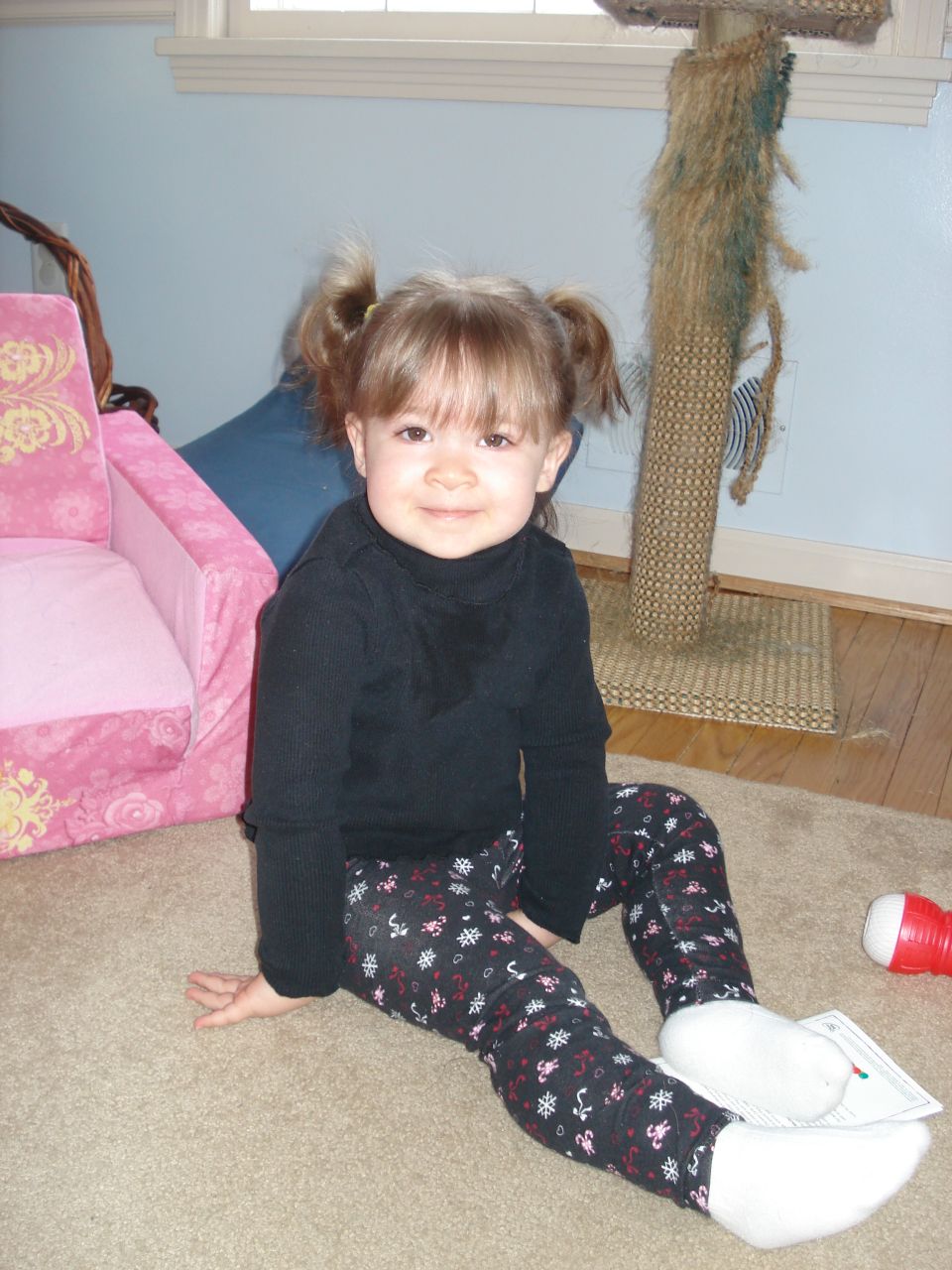 Young girl with pigtails sitting on floor next to pink child's chair and cat tree.