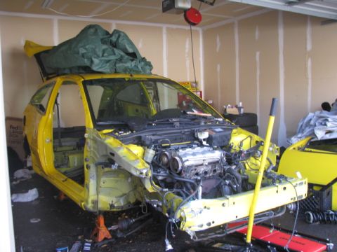 2003 Ford Focus - A yellow hatchback is undergoing disassembly in a garage, with its engine exposed and parts removed.