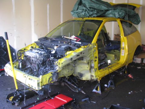 2003 Ford Focus - Disassembled yellow car, likely a Ford Focus, in a garage with tools and a tarp.