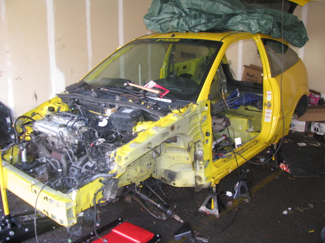 Yellow car with front end removed, engine exposed, doors open, and interior stripped. Car is on jack stands in a garage