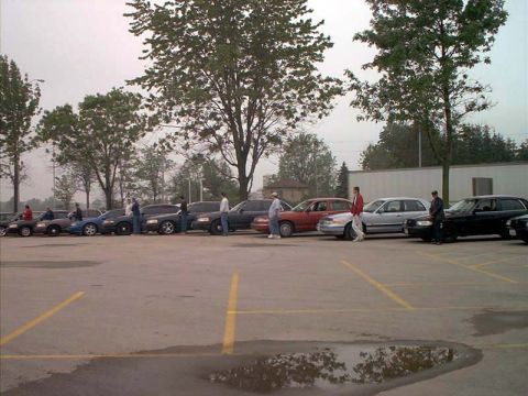 2000 Ford Crown Victoria - line of sedans and SUVs parked diagonally in an asphalt lot with several people standing between them on an overcast day.