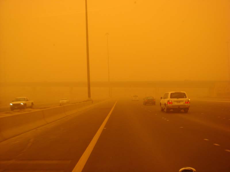 Vehicles driving on a highway obscured by a sandstorm, with a white SUV visible in the foreground