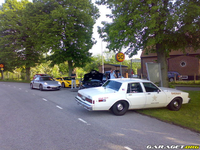 White sheriff car parked on grass, side view. Other sports cars visible