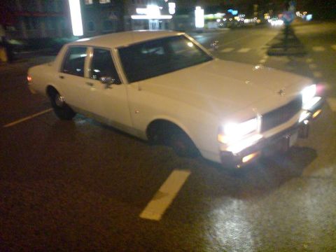 1998 Ford Crown Victoria - A white sedan is parked on a wet street at night with its headlights illuminated.
