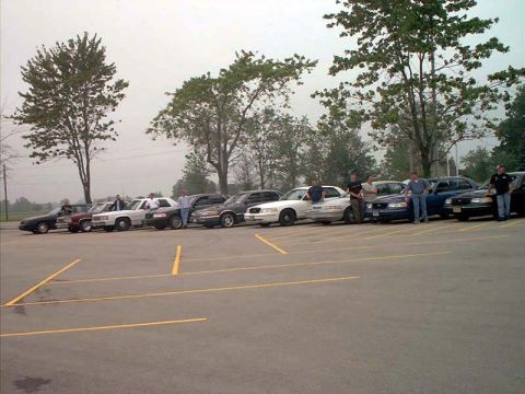 2000 Ford Crown Victoria - A lineup of Ford Crown Victoria police cars and civilian sedans in a parking lot.