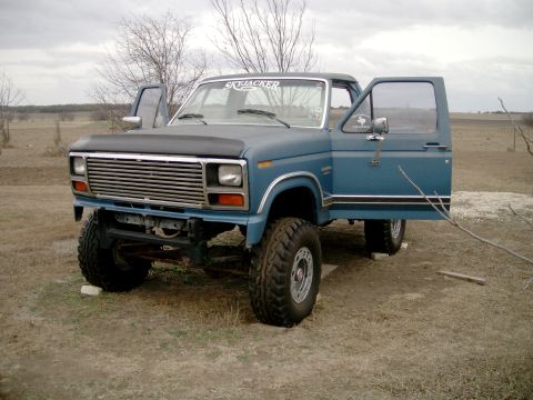 1984 Ford Bronco - A blue Ford Bronco sits in a field with its doors open, showing off its lifted suspension and aftermarket grille.